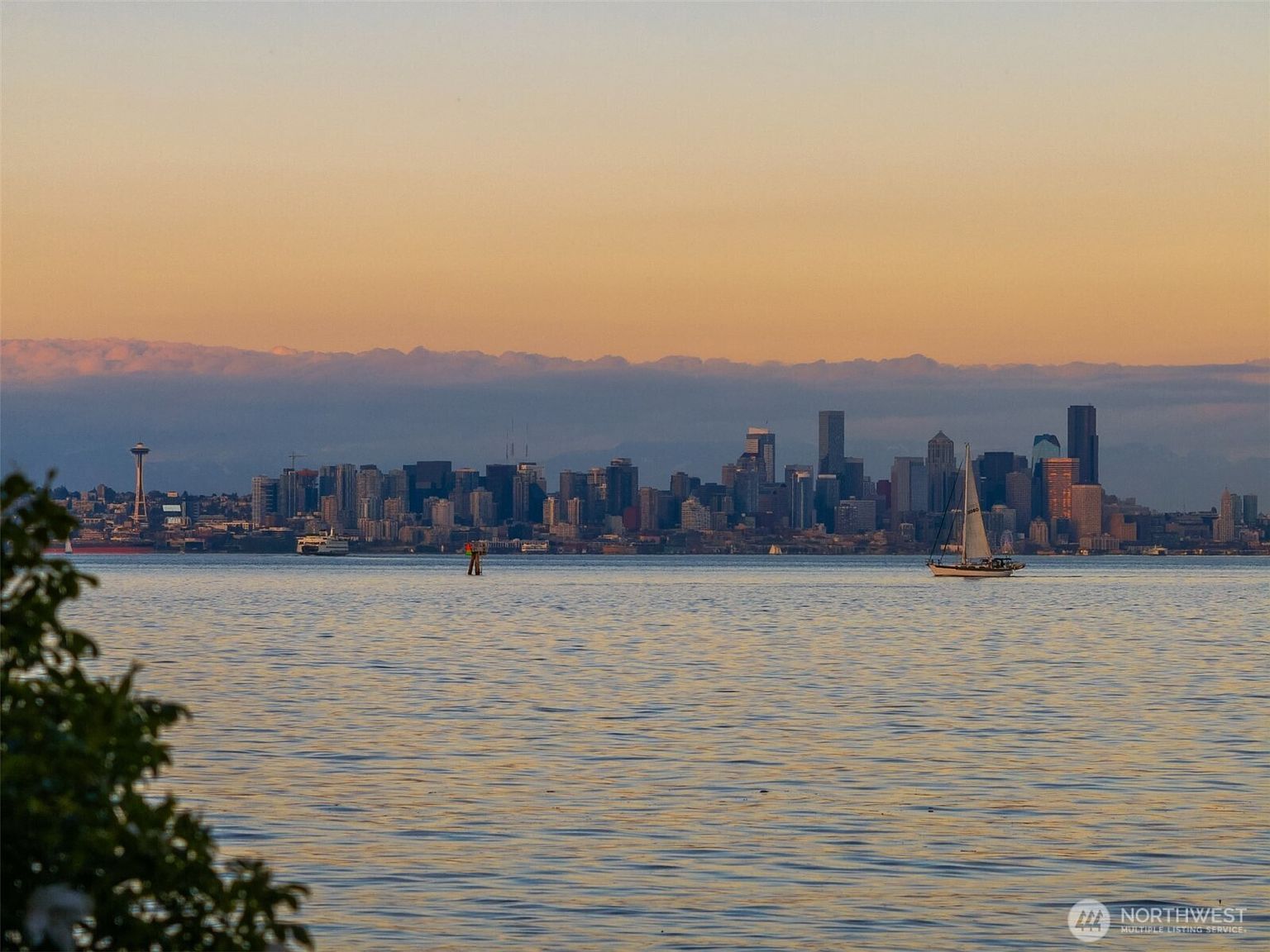 The view east from Rockaway Beach: a sailboat passing the Seattle skyline at sunset, Mount Baker shadowed in the distance.