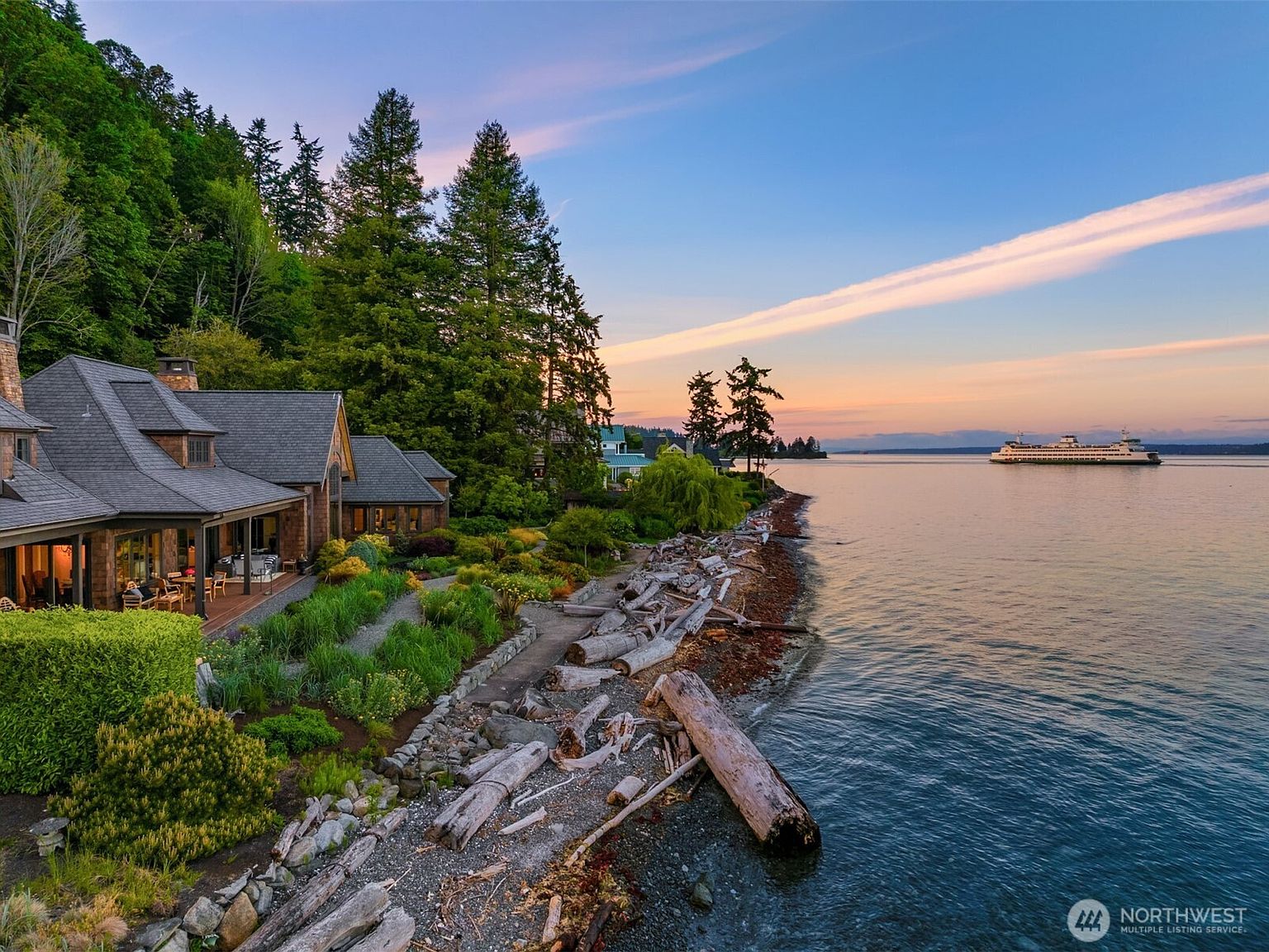 Twilight aerial of the 5128 Rockaway Beach Road shingle-style estate built into the forested shoreline of Bainbridge Island, with a Washington State Ferry crossing Puget Sound at dusk.
